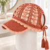 Terracotta crochet visor cap with pale peach shell trim and tassels displayed on a white stand